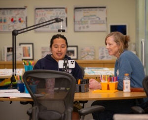 Ryenchindorj Erkhembayar sits at table in front of recording records. Sue Goldie, also seated at table, observing and providing mentorship, smiles off-camera