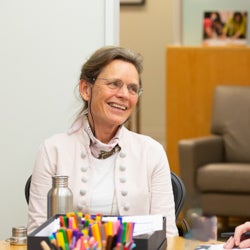 Myriam Hunink, wearing glasses and a business blazer and smiling off-camera, is sitting at table with colorful markers in an office setting.