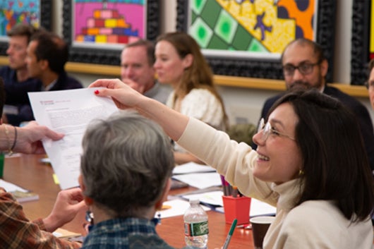 Student seated at conference room table with others, handing paper to someone off camera while smiling
